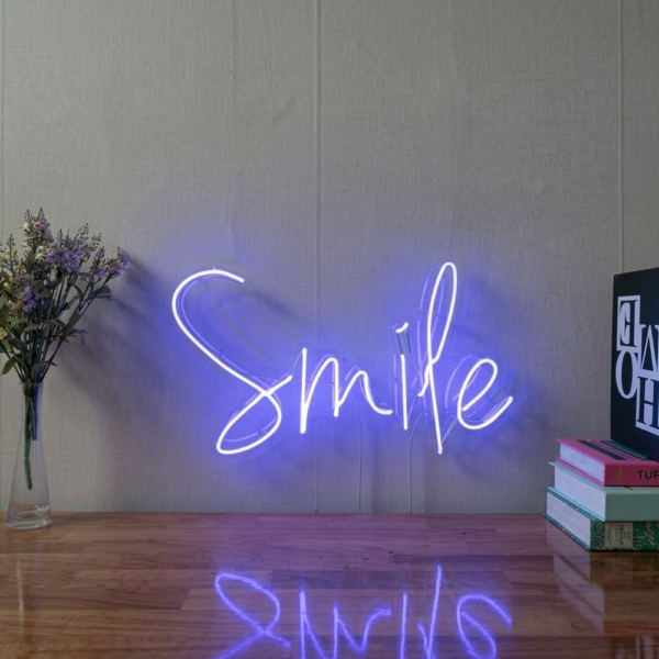 El yazısı Smile cam neon tabela, gri duvar önünde mor tonlu ışık ve ahşap masa üzerindeki yansıması