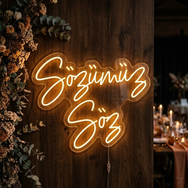 Koyu ahşap yüzeyde çiçeklerin yanında sıcak sarı ışıkla iki satır Sözümüz Söz yazılı LED neon tabela